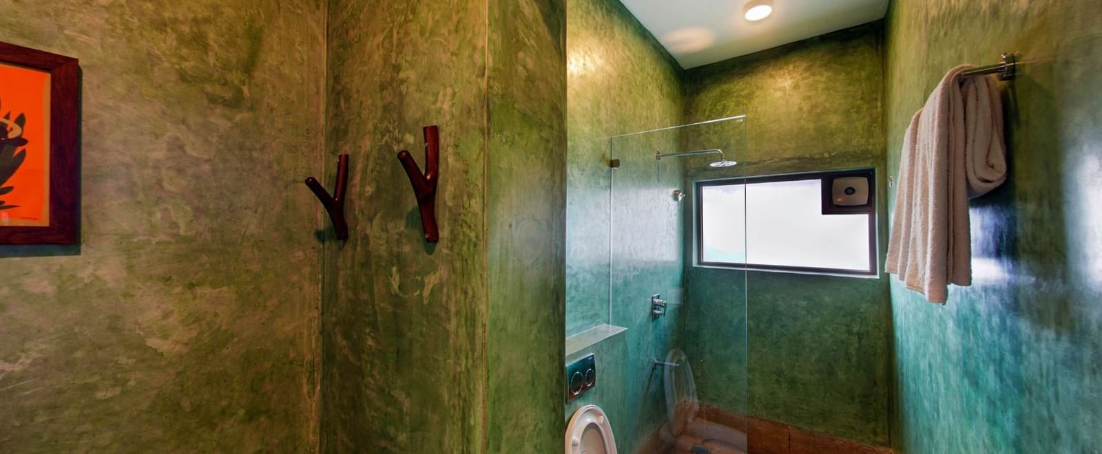 Narrow green-tiled bathroom with wall hooks, toilet, ventilator, and ceiling lights at Fireflies Resorts, Kabini.