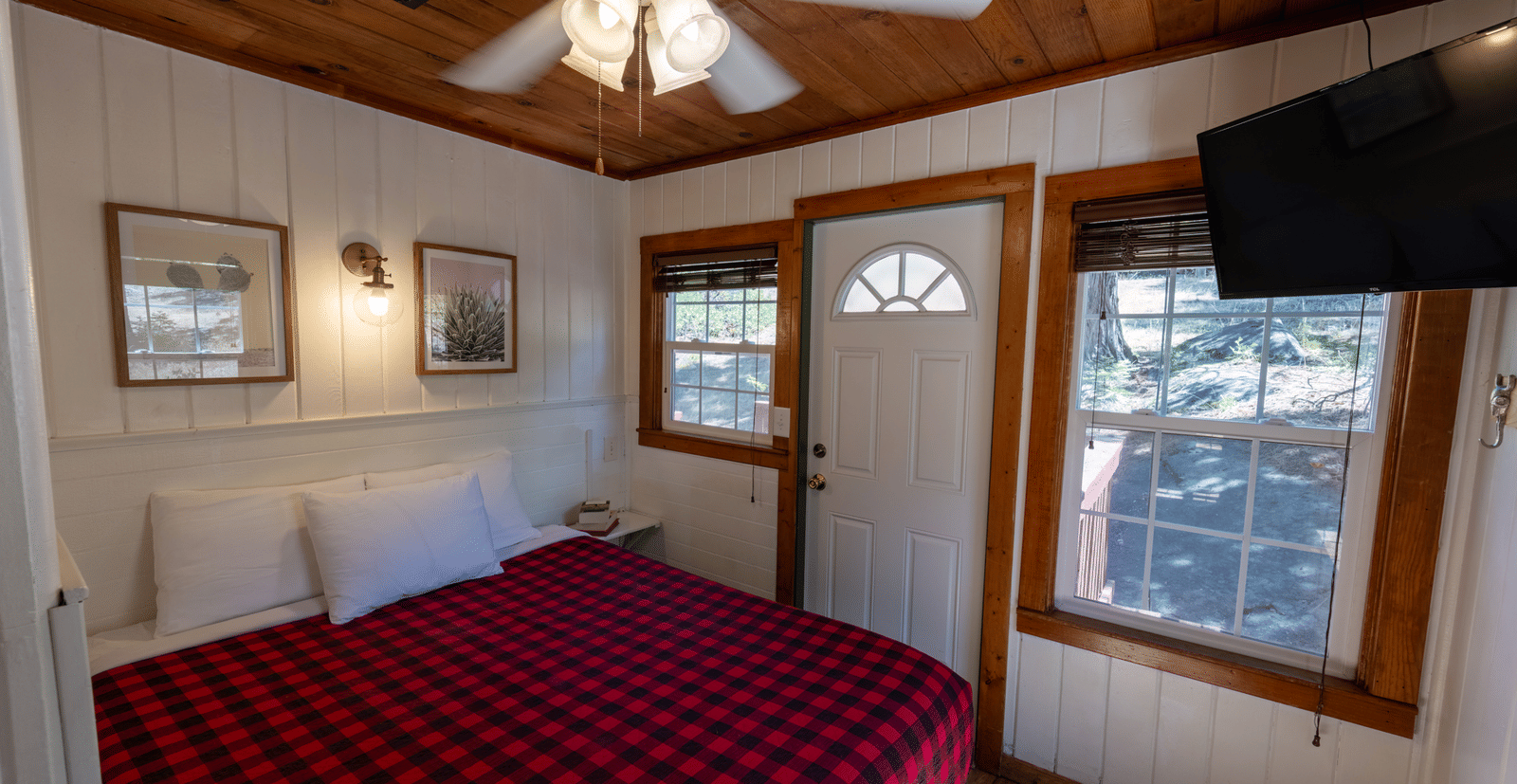 A cozy cabin bedroom with wood-paneled walls and ceiling, a large bed with a red and black checkered comforter, and a wall-mounted TV - Shaver Lake Village Hotel