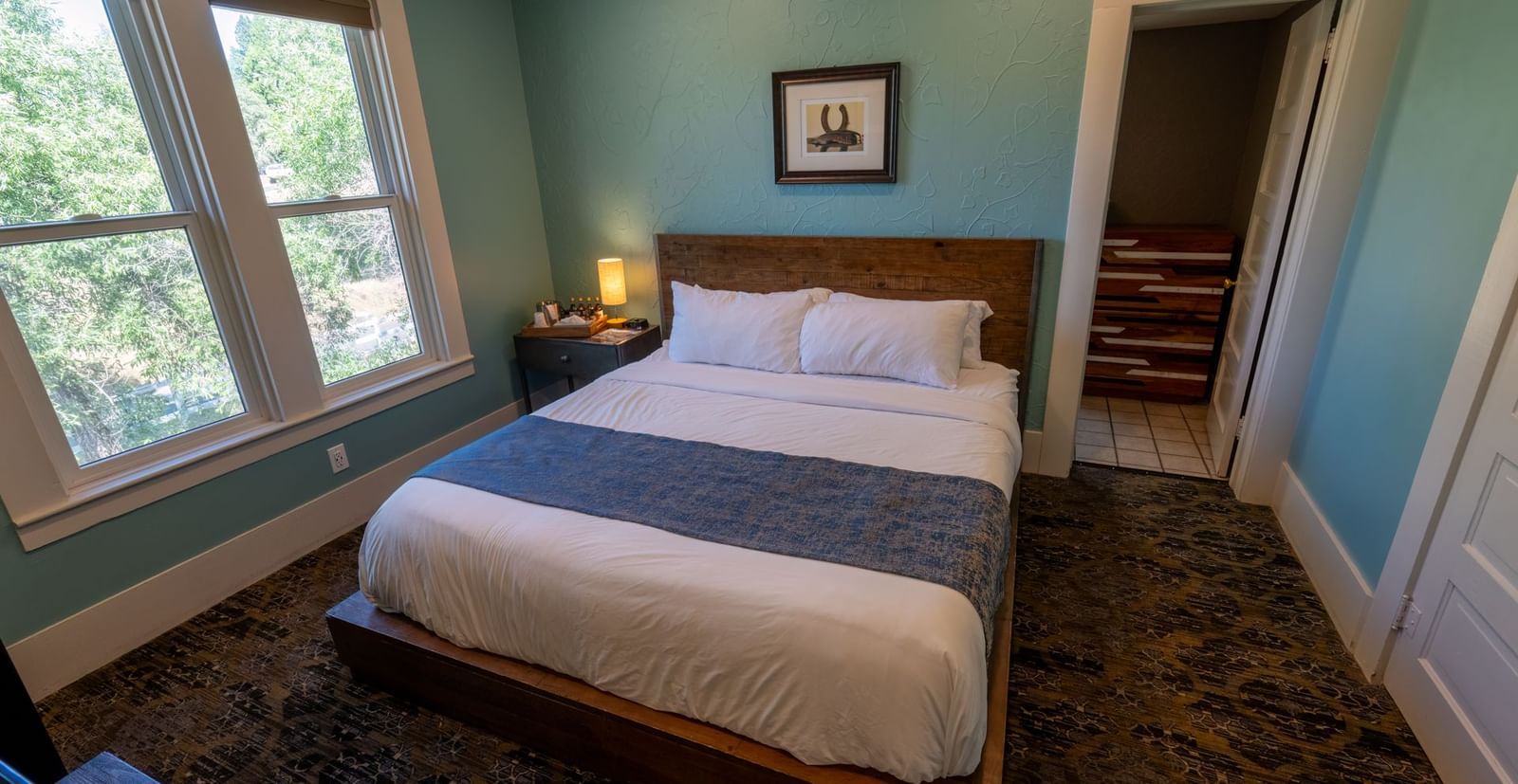 Corner view of a Family Suite at The Groveland Hotel featuring a plush bed, carpeted flooring, and a wood-framed glass window offering a view of the greenery nearby.