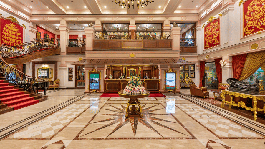 Grand and opulent lobby of Mayfair Lagoon, Bhubaneswar, featuring a grand chandelier, intricate marble flooring, ornate golden decor, vibrant red banners, and a majestic double-staircase