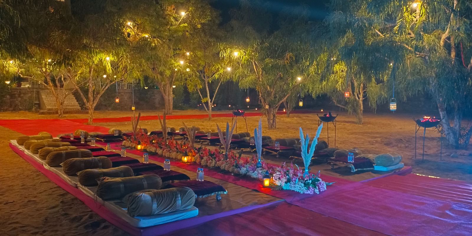 An outdoor area at night at the Bamboo Saa Mulberry Resort, Pushkar, with seating arrangement on the ground illuminated by string lights.