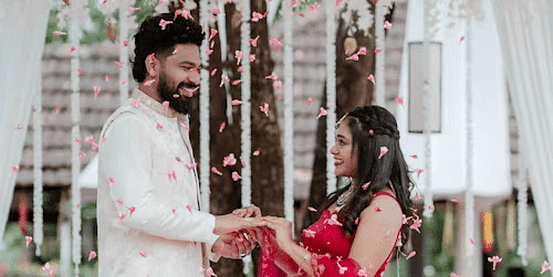 Romantic proposal moment under floral decor, with groom kneeling before bride in red bridal attire.