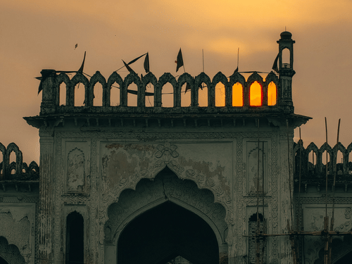 Silhouetted ornate stone archway and parapet against a warm, golden sunset sky with soft, glowing clouds behind it.