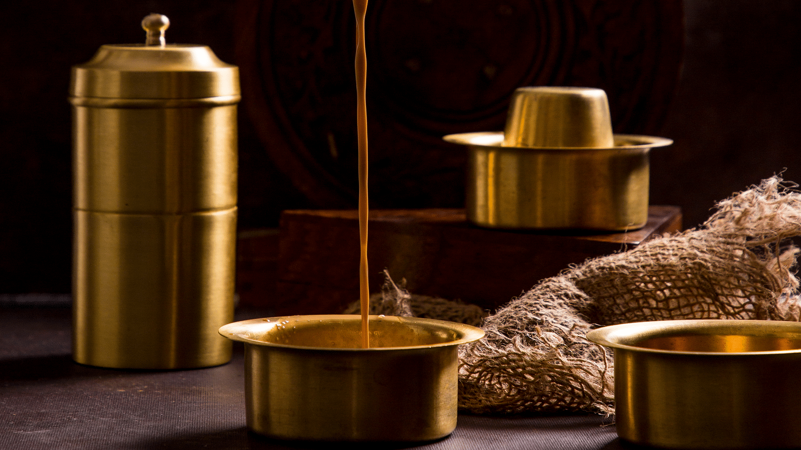 A hand pouring tea into a brass vessel surrounded by similar vessels and a container | Kamats India