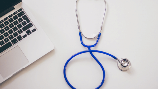A blue stethoscope is placed on a white surface next to the corner of an open laptop.