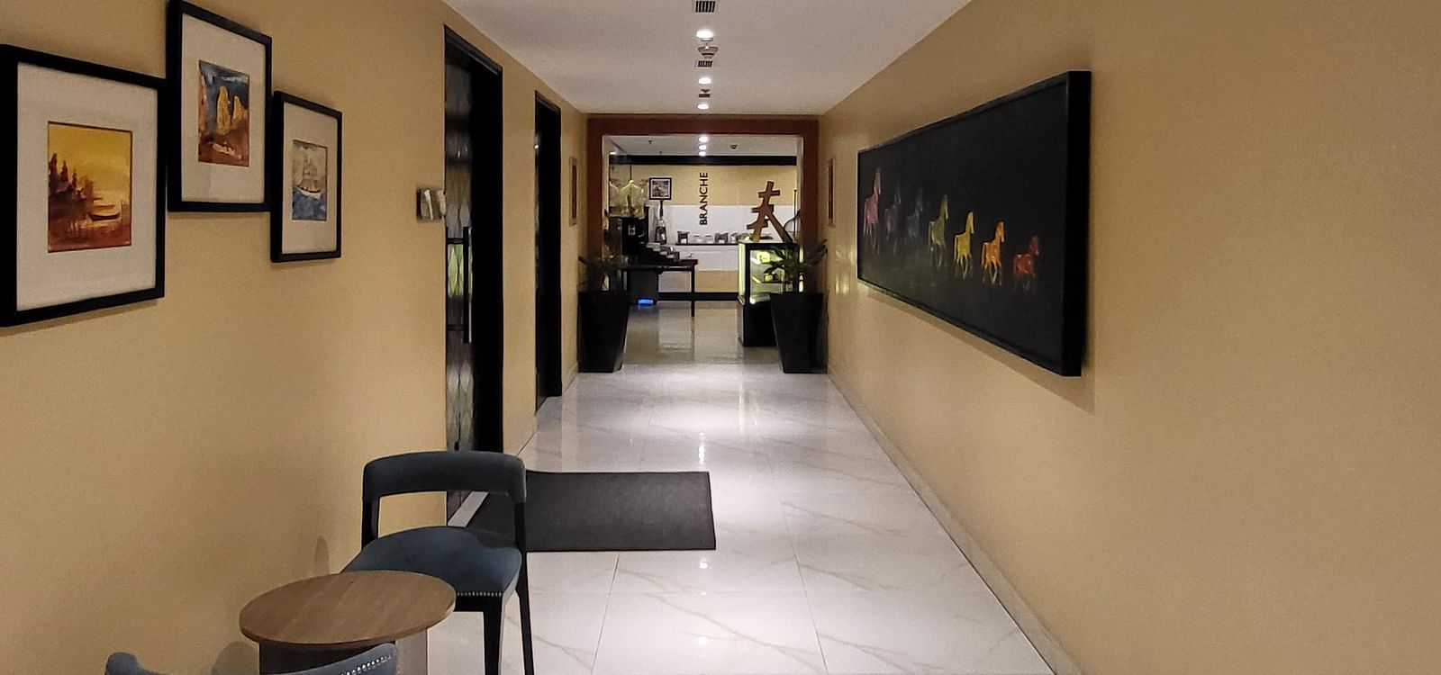 An image of a corridor at Golden Suites Gurgaon featuring seating small chairs and tables at one side, photo frames on the walls, and several doors leading to each room