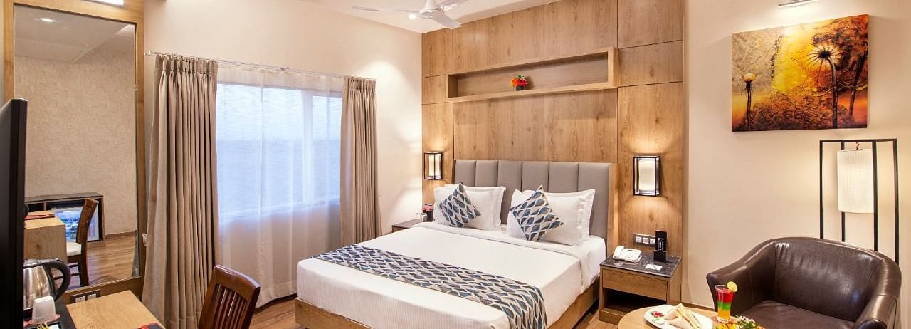 A hotel room showcasing a double bed with white and grey bedding, a wooden bedside table with a lamp, a work desk area with a chair and television, and a small round table with a brown armchair, complemented by wooden flooring at Lords Inn Motikhavdi