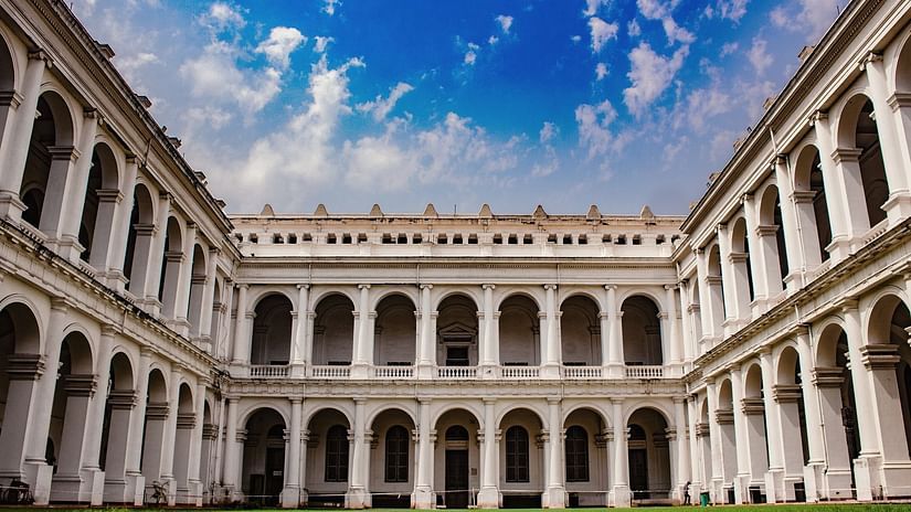 A grand white colonial-style building with arched corridors surrounding a green courtyard under a vibrant blue sky with scattered clouds.