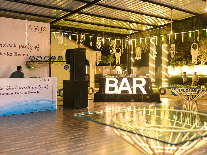 An event venue featuring a bar counter with neatly arranged bottles, glass table setups, and warm lights hanging from the ceiling for decoration | VITS Daman Devka Beach