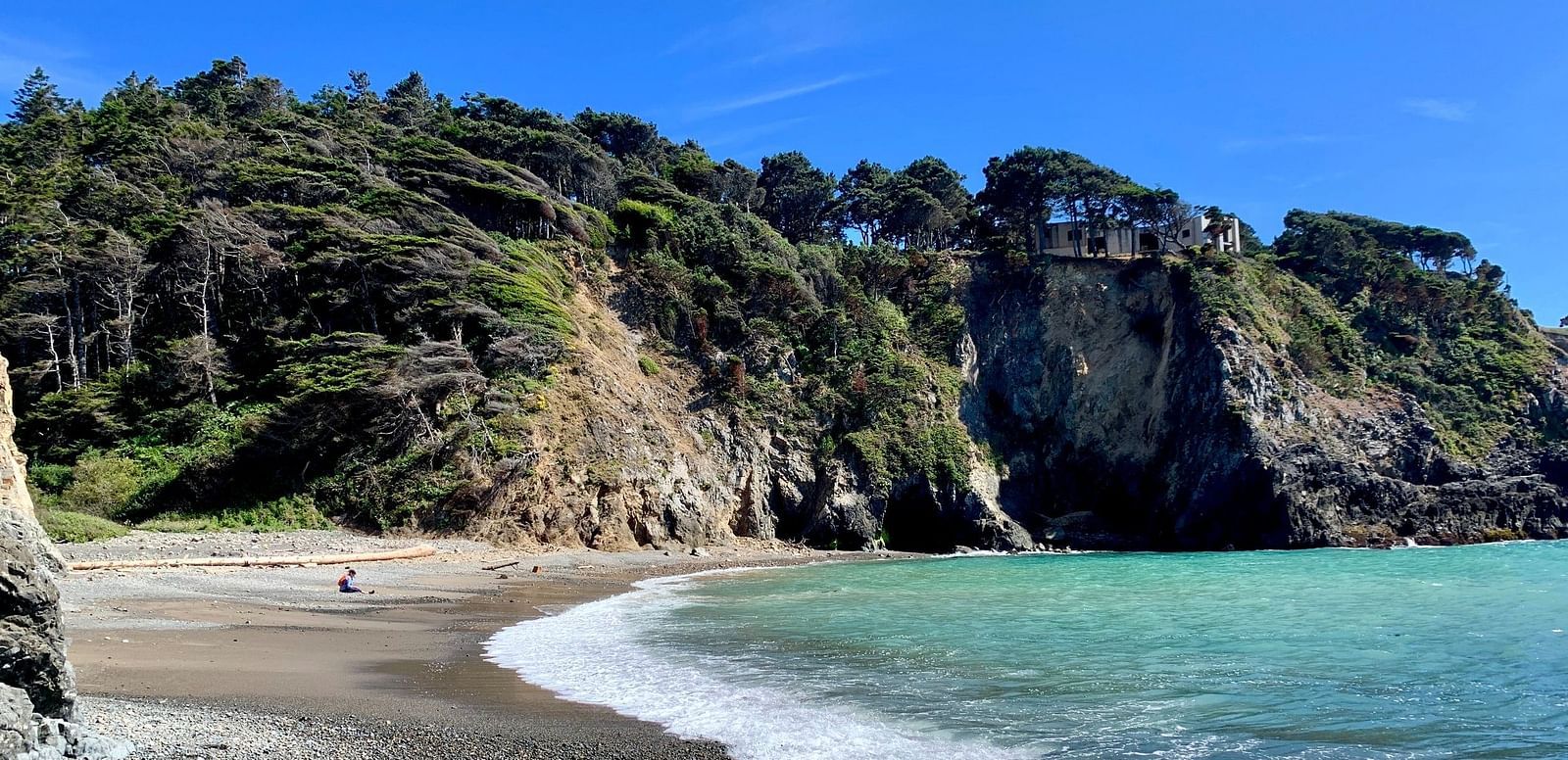 A curved shoreline meets a cliffside covered in vegetation, with a structure perched atop the rocks under a clear sky.