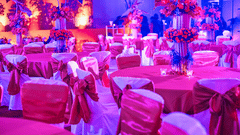 A brightly lit banquet hall is decorated in pink and red tones for a celebration, with tables and chairs set up for guests.