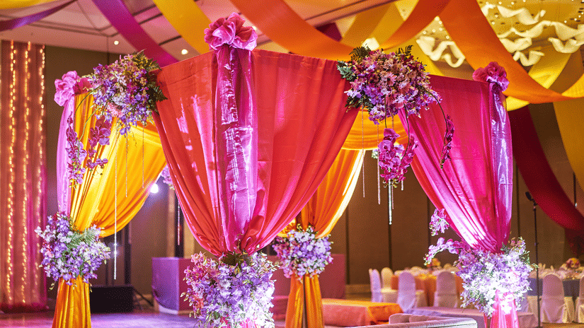A colourful, decorated wedding venue is set up with bright orange and pink fabrics draped over a structure, creating a festive atmosphere.