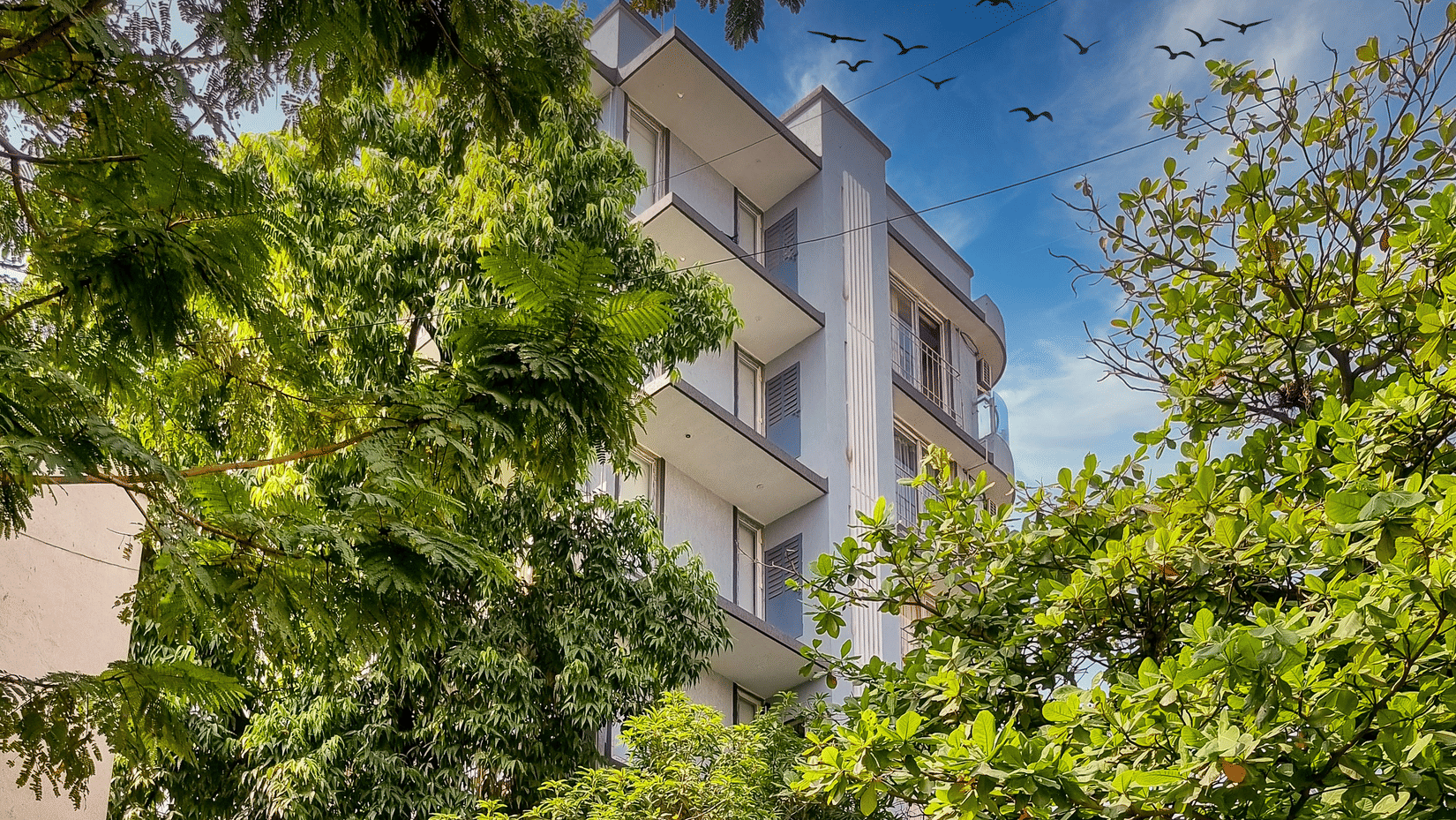 The building exterior of Home2 Residency, Mumbai Airport, with multiple floors, surrounded by trees, tiled walkway in foreground, boundary wall with pattern, and potted plants near entrance.
