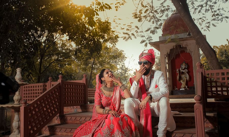 A newlywed couple in wedding attire sits on temple stairs, laughing and playfully covering their mouth at a Jaipur's Top Wedding Resort - Umaid Palace - A Lakeside Heritage Palace Resort