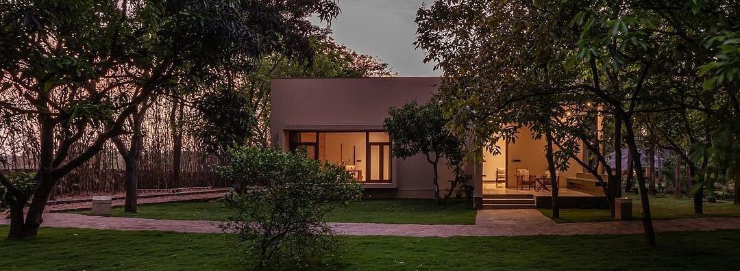 An exterior view of a building with lit windows, surrounded by trees and greenery at dusk - Shreyas Retreat