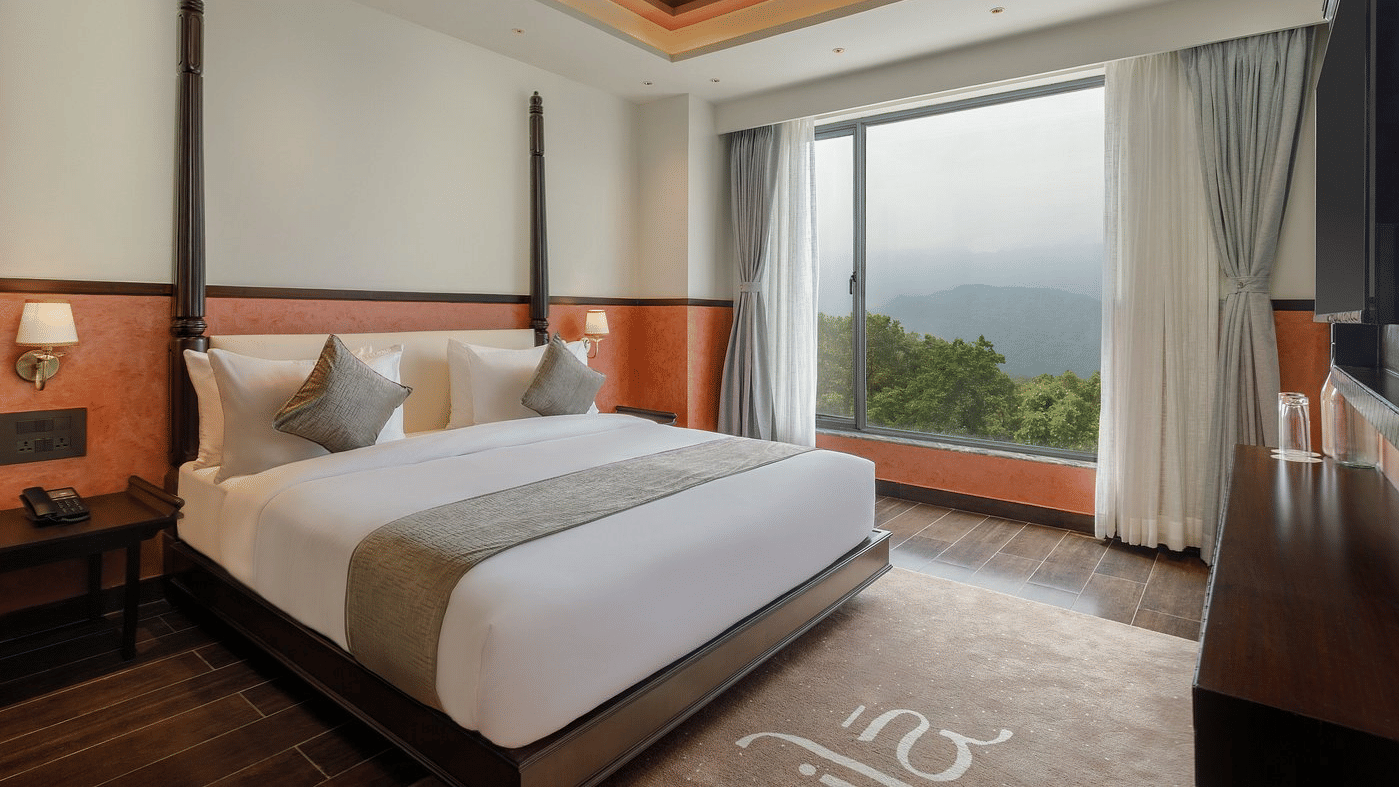 Guest room with a four-poster double bed, terracotta accent walls, a coffered ceiling and a wide window facing wooded hills at Ila Resort, Maulakali.