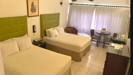 A hotel room showcases two beds with green headboards, a dresser, and a television, with a ceiling fan visible | Executive Double Room | Altamont Court, Jamaica
