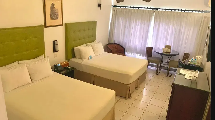 A hotel room showcases two beds with green headboards, a dresser, and a television, with a ceiling fan visible | Executive Double Room | Altamont Court, Jamaica
