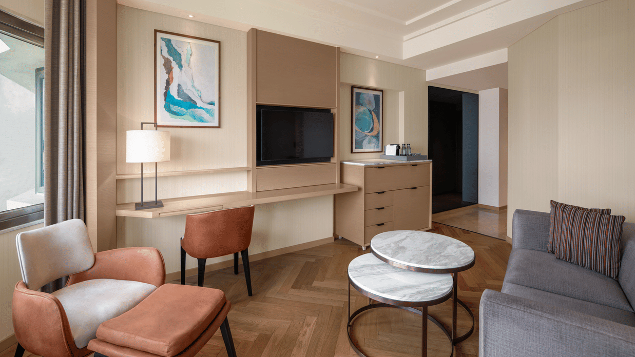 Inviting living area of a suite room at The Residency Chennai, featuring a large cozy sofa, a comfortable chair, a round table, and a wall-mounted TV.