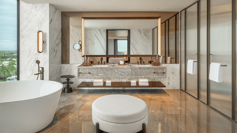 Presidential Suite bathroom with a freestanding bathtub, a marble double vanity, a round ottoman and fluted-glass doors at The Residency Signature Madurai.