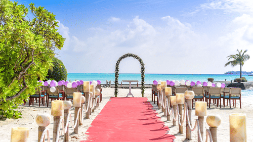 Scenic beach wedding venue with a red carpet aisle leading to a floral arch, elegant guest seating, decorative candle-lined pathway, and breathtaking ocean views under a bright blue sky.