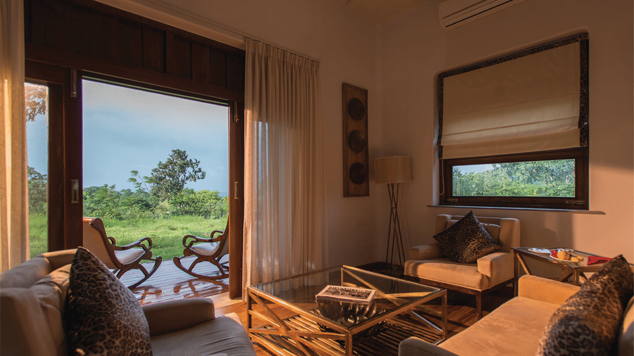 Sun illuminating the interiors of a room at The Serai Bandipur featuring a sofa set and a patio seen through the doorway with a couple of chairs facing the trees outside