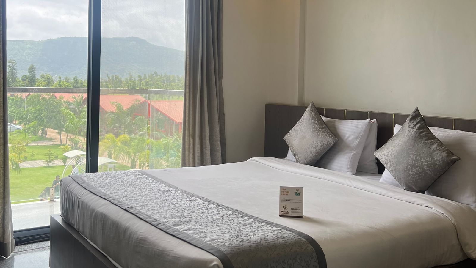 Close up of the queen size bed in the Daksh Bliss room at Daksh The Valley Resort, Saputara, with curtains drawn, offering views of the greenery surrounding the room.
