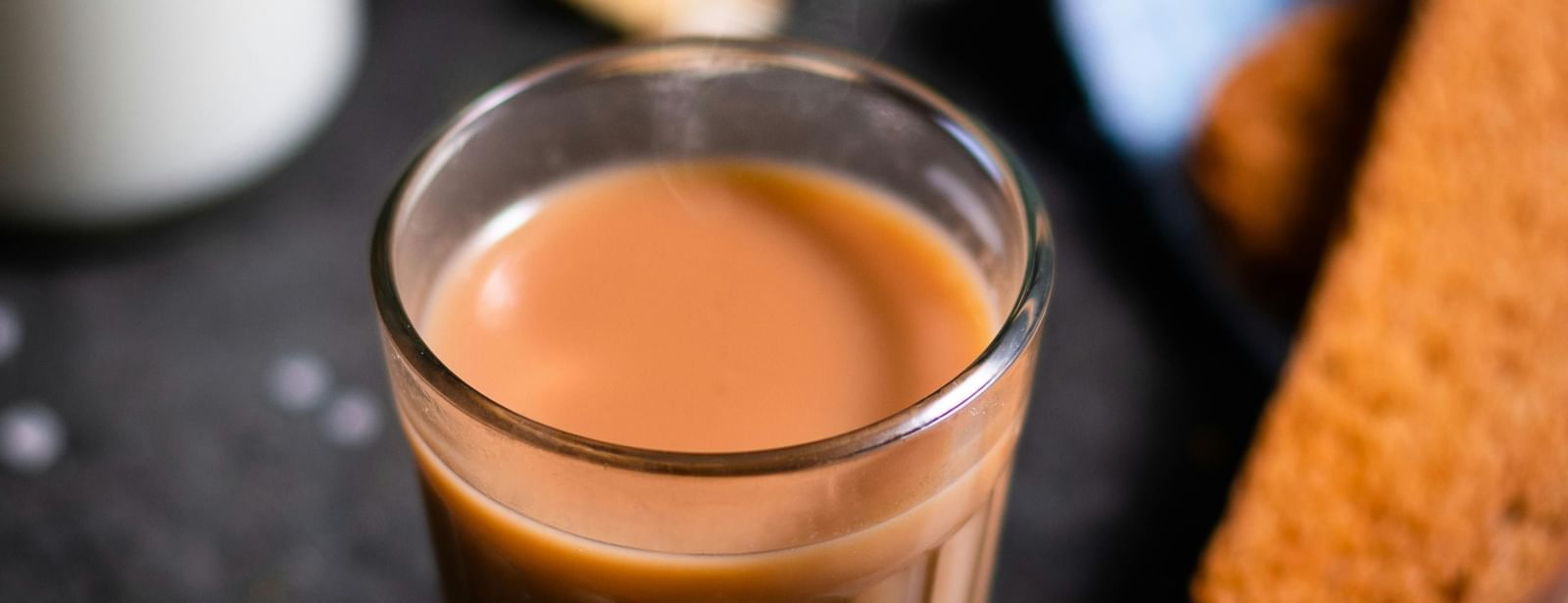 A glass of tea placed on a black surgace with a spoon and a piece of ginger next to it.