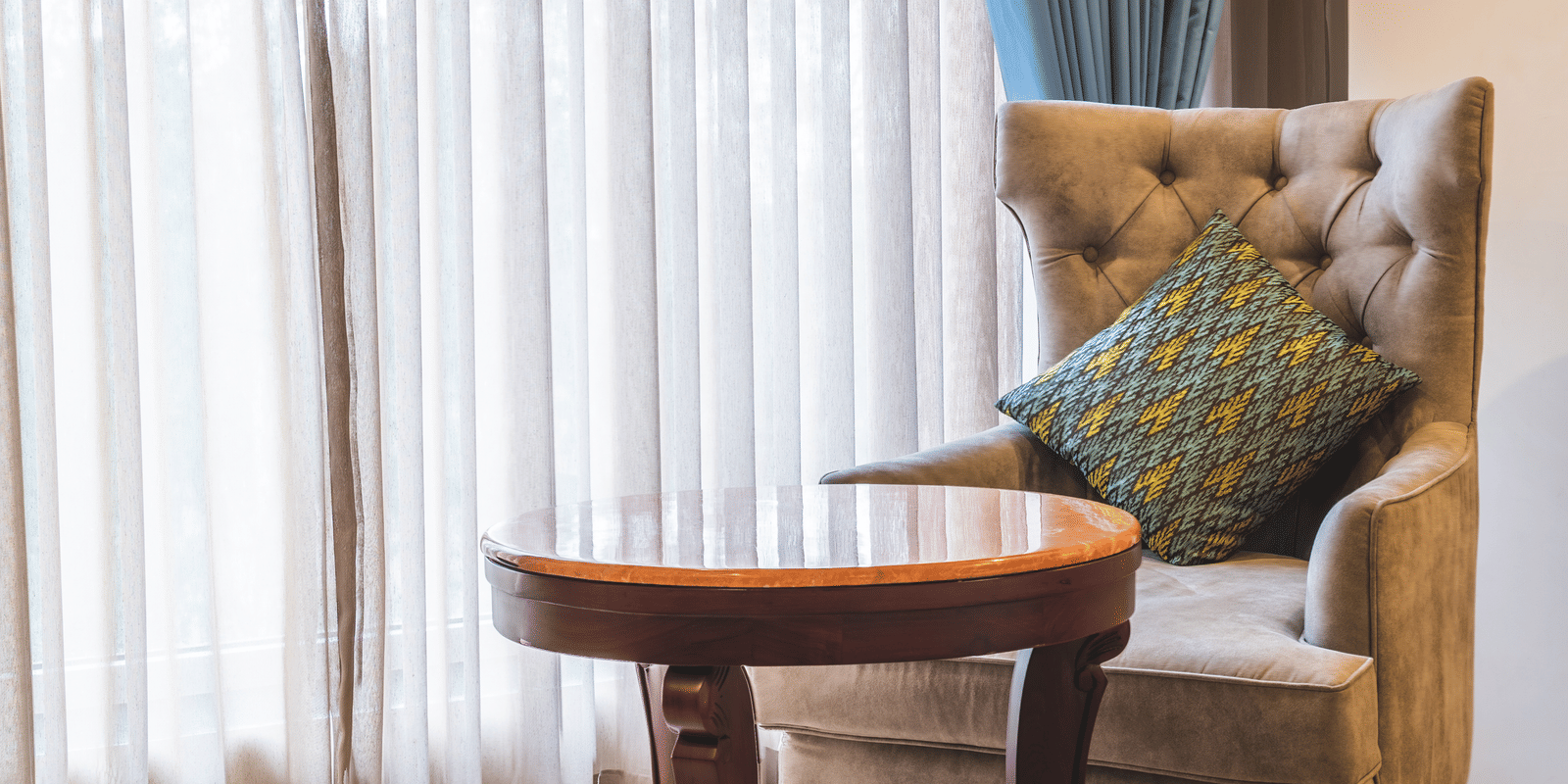Comfortable tufted armchair with a cushion next to a small round wooden table, set against a window with sheer curtains at Hotel Hukam's Lalit Mahal.