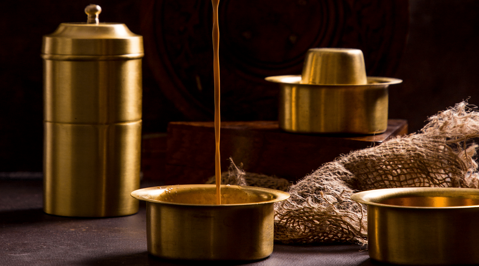 A hand pouring tea into a brass vessel surrounded by similar vessels and a container | Kamats India
