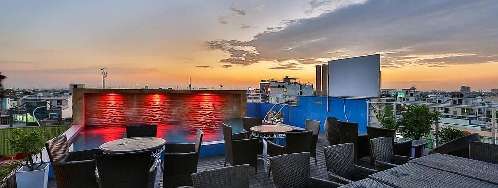 A rooftop terrace during sunset with wicker furniture, a lit pool and a view of the city skyline - TGI Apple Inn, Jaipur 