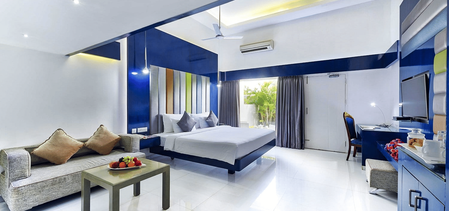 A hotel room in ECR with blue walls, a double bed, a seating space with sofa and a chair, and the window opening to the greenery at Grande Bay Resort & Spa, Mamallapuram.