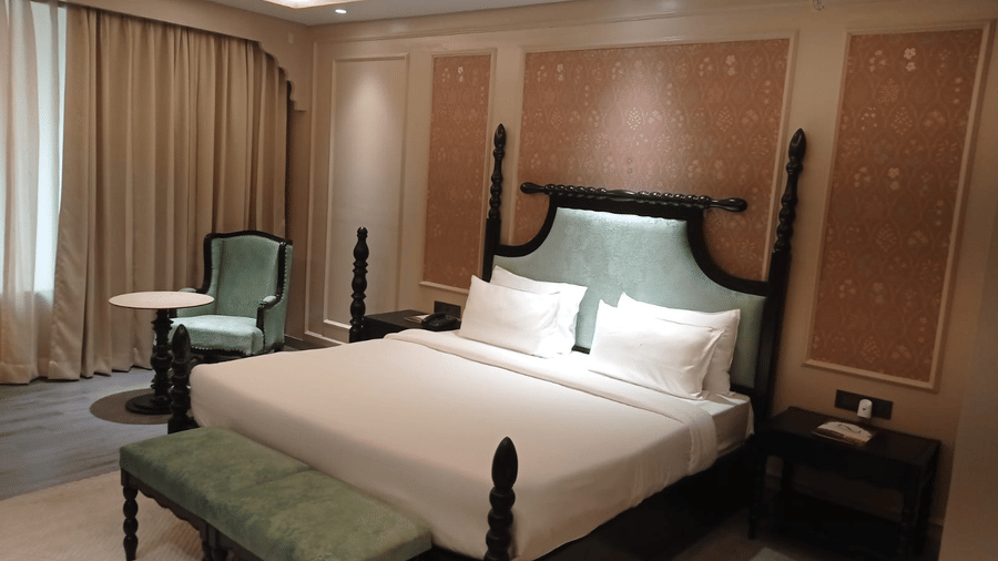 A bedroom with king size bed, side tables, chair, bench and window curtain at Beelwa Palace, Jaipur.