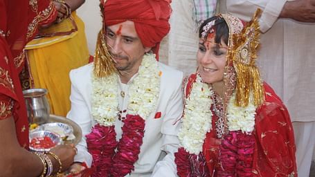 A couple dressed for a wedding ceremony, wearing garlands and traditional attire, is the focus as someone offers a plate.