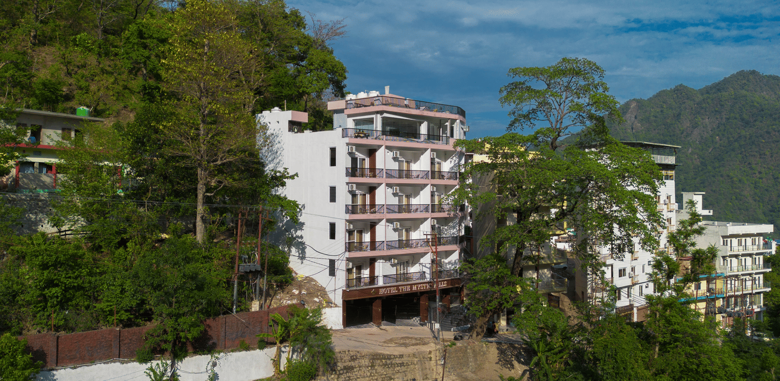 An exterior of a building with multiple storeys, balconies, and windows, surrounded by trees on a hillside in Perfectstayz Premium Mystic Falls, Rishikesh.