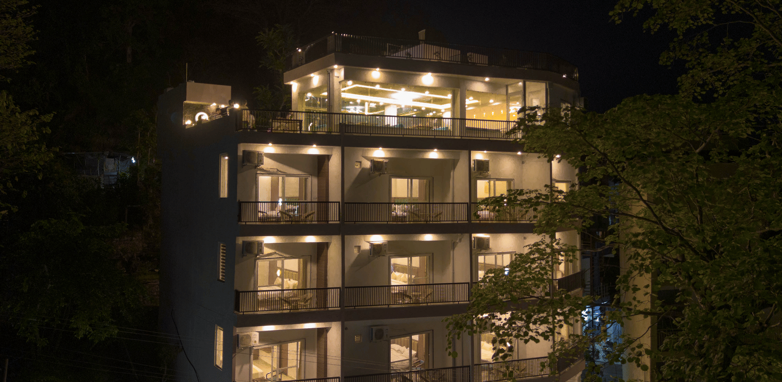 A multi-storey building exterior at night, with windows lit up and trees surrounding the structure in Perfectstayz Premium Mystic Falls, Rishikesh.