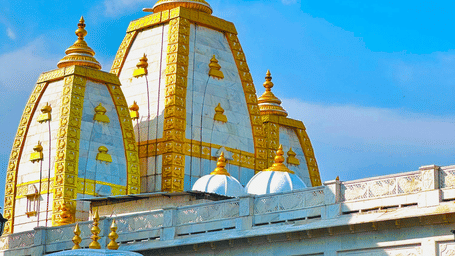 Shining golden domes of a temple rise above white arches, topped with a red flag against a brilliant blue sky.