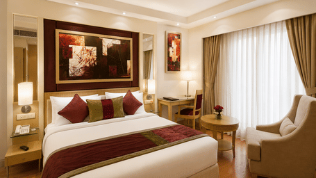 Hotel room with a bed dressed in white and maroon linen, framed wall artwork, a beige armchair and long green curtains by the window nearby.