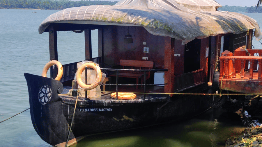 A traditional wooden houseboat anchored on a calm river with a thatched roof and open deck seating at Paradise Lagoon Resort, Udupi.