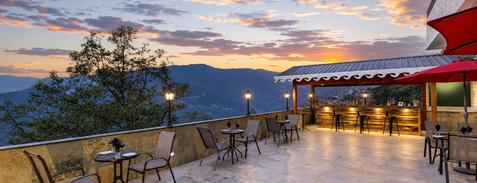 Terrace bar at Valmere Himalayan Hotel & Spa, Gangtok at sunset, with 3 wooden barstools, patio seating, red umbrellas, and mountain views beyond the railing.