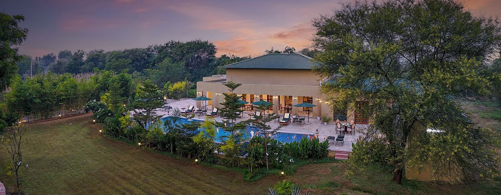 An elevated shot of a luxurious resort property featuring a winding swimming pool, surrounded by trees and manicured lawns, under a colourful evening sky - Singhvilas, Ranthambore