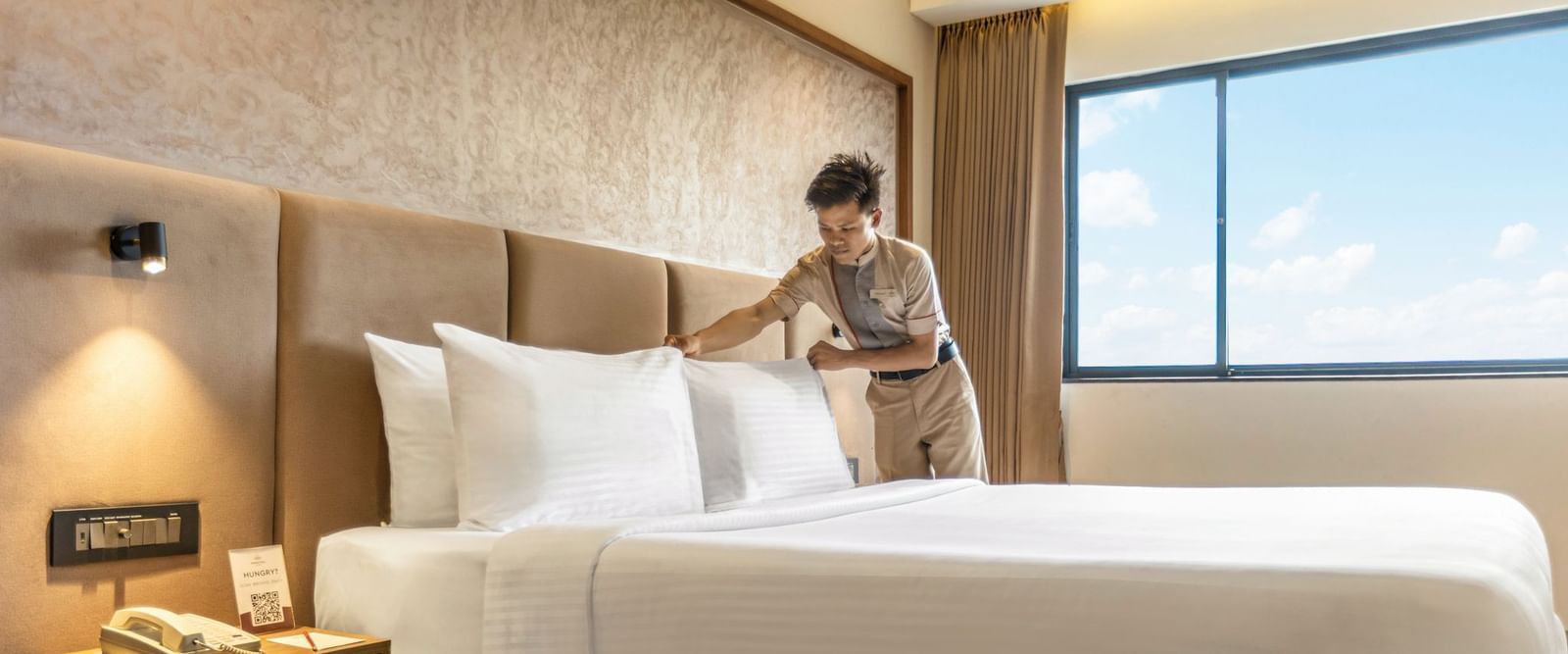 A person arranging pillows on a bed in a room at Mango Hill Central, Chennai. There is a large window on the far wall.