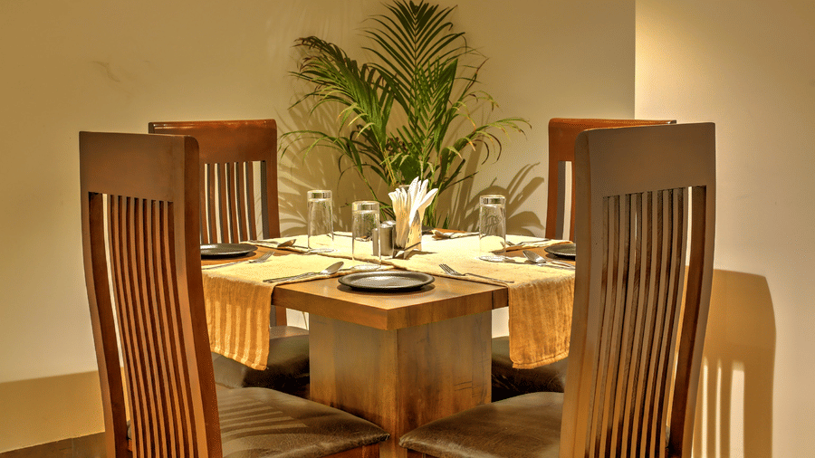 A close-up of a square wooden dining table for four, neatly set with yellow runners and modern tableware near a green plant - Hotel 91 HUDA City Centre, Gurugram