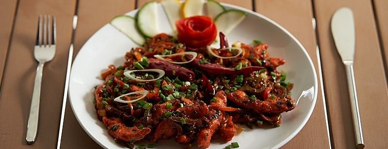 A white plate of  vegetables garnished with fresh herbs and a carved tomato rose, served with sliced cucumber and lime, placed on a wooden table with a fork and knife at The Hamuse Hotel, Kodaikanal.