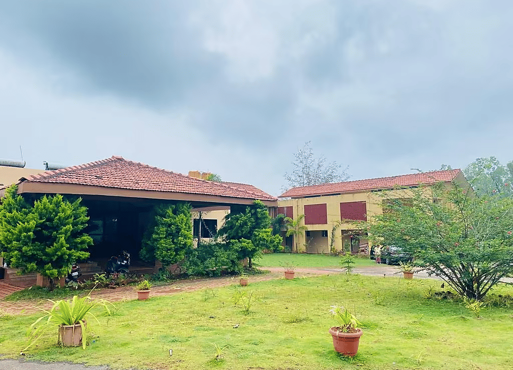 Building with tiled roof and green plants in front, surrounded by grass and potted plants on a cloudy day at  Paradise Wild Hills Resort, Kollur.