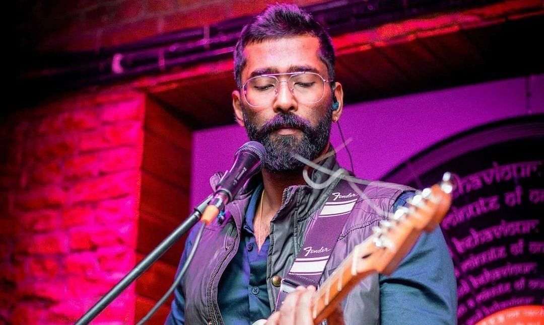 A bearded musician wearing glasses and a stylish vest passionately plays a white electric guitar on stage. 