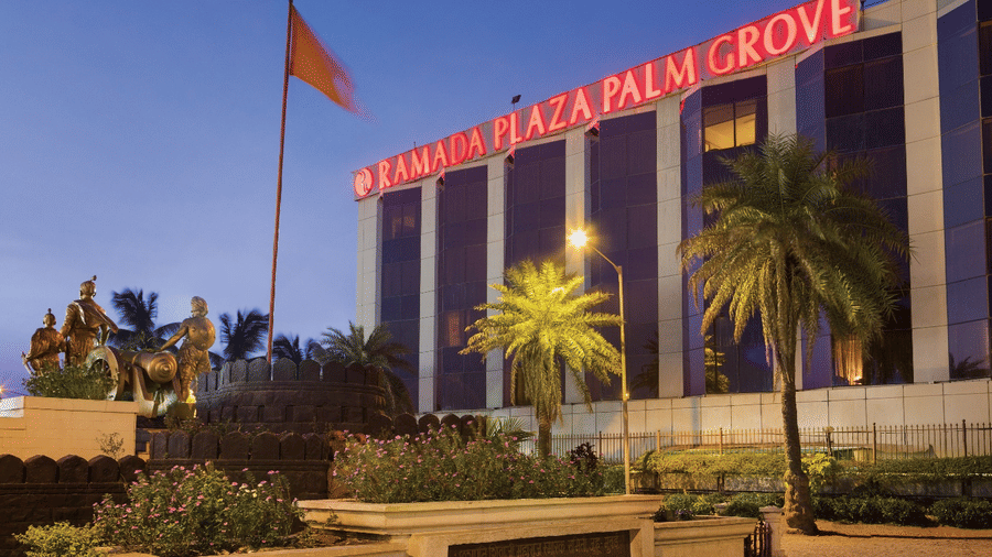 Exterior view of Ramada Plaza by Wyndham Palm Grove, showcasing its illuminated facade, palm-lined surroundings, and prominent city landmark setting.