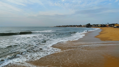 waves crashing on an empty beach with trees & buildings on the far side and white clouds on blue sky in the background