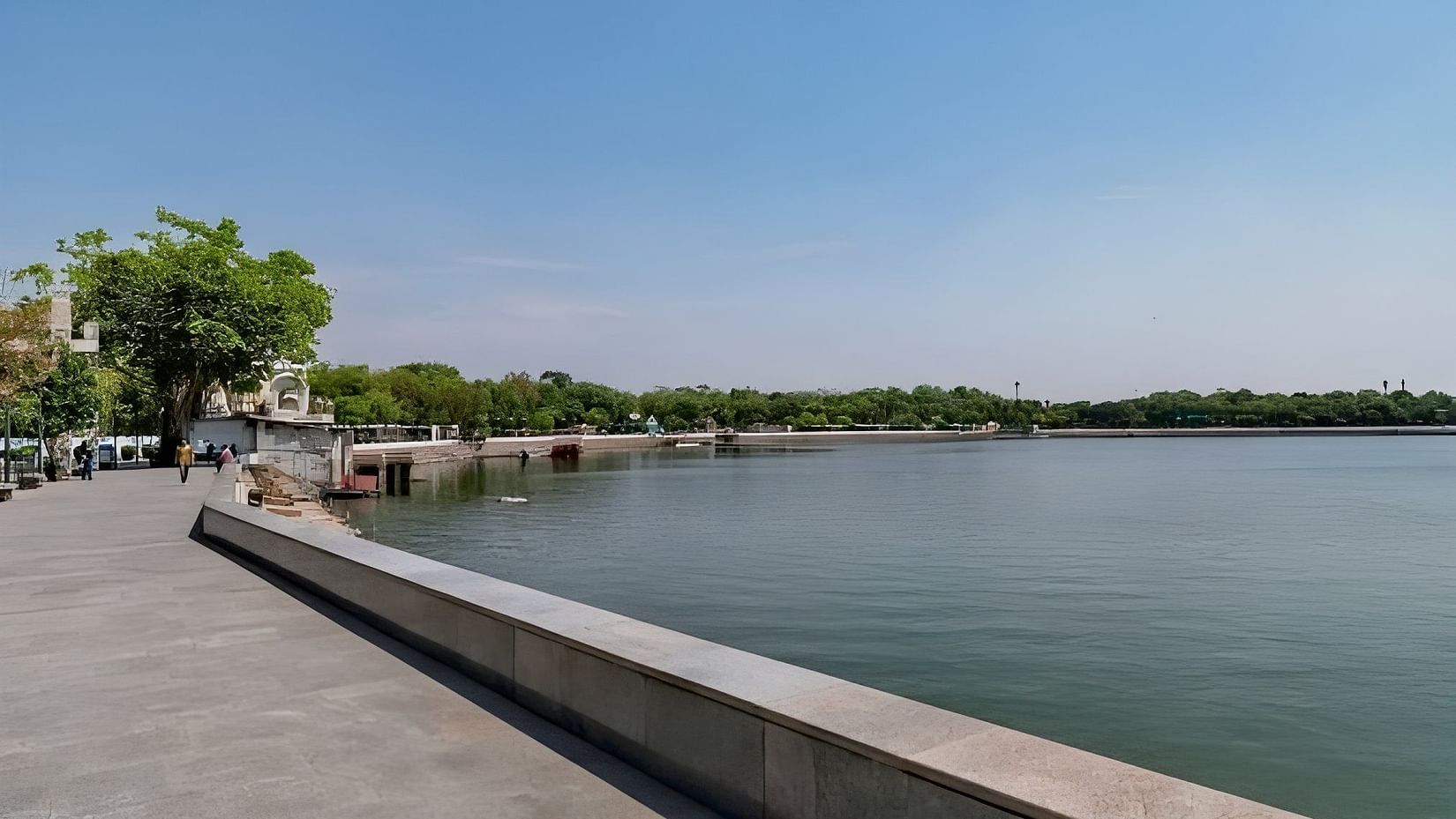 A peaceful daytime view of a lakeside promenade with a wide walking path, calm water, and greenery in the distance under a clear blue sky.