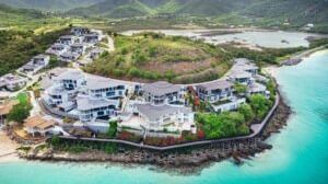 An aerial view of a coastal development with numerous white buildings nestled along a vibrant blue bay surrounded by green hills.